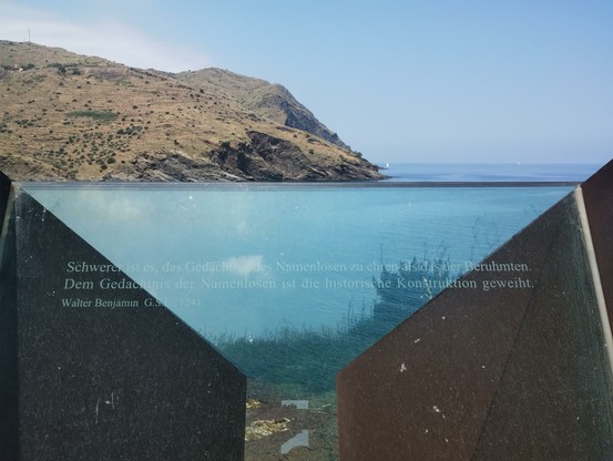 Blick auf eine Glasscheibe mit der Aufschrift:
»Schwerer ist es, das Gedächtnis der Namenlosen zu ehren als das der Berühmten.
Dem Gedächtnis der Namenlosen ist die historische Konstruktion geweiht.
Walter Benjamin, G.S. I (auf der Glasscheibe als L), 1241«
Im Hintergrund Meer und eine Küste