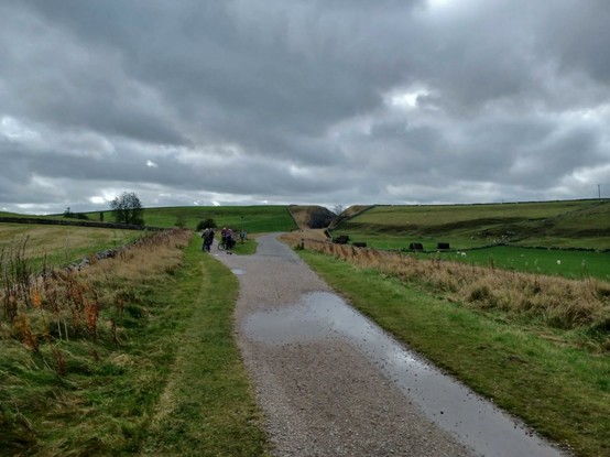 The Tissington Trail outside Parsley Hay. The countryside is green and hilly, the clouds are grey and threatening. The picture shows a fork in the trail. The right hand branch goes to Ashbourne, the left hand branch is the High Peak Trail to High Peak Junction. There are a couple of cyclists about to head onto the High Peak Trail.