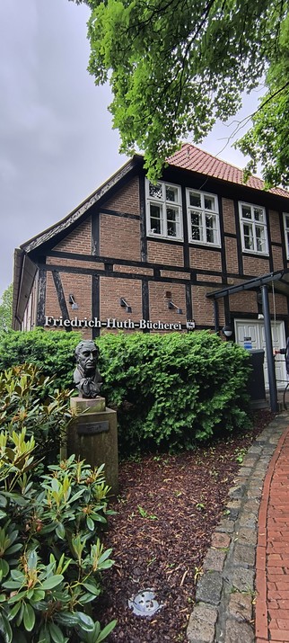 Buitenkant van de bibliotheek, met borstbeeld van de persoon waarnaar deze vernoemd is: Friedrich Huth Bücherei. Vakwerk met baksteen en veel groen ervoor.