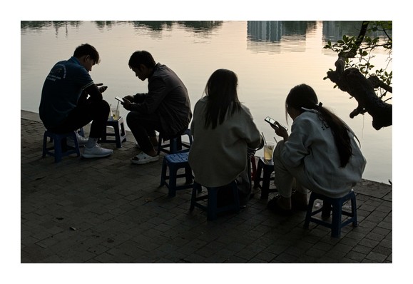 The image depicts a serene scene by a body of water, likely a lake or pond, during what appears to be the late afternoon or early evening. Four individuals are seated on small, colorful plastic stools arranged in a semi-circle. They are all engaged with their smartphones, suggesting a moment of leisure or relaxation.

The individuals are silhouetted against the soft light reflecting off the water, creating a calm and peaceful atmosphere. Two of the individuals have drinks placed beside them on the stools, possibly enjoying a casual beverage. The ground is paved with bricks, and there is a tree branch extending into the frame from the right side, adding a natural element to the scene. The overall mood of the image is tranquil and reflective.