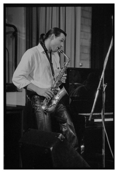 Black and white photograph of a saxophone player. He is seen from the side looking to the right. It looks as if he is communicating with somebody just outside the frame.
