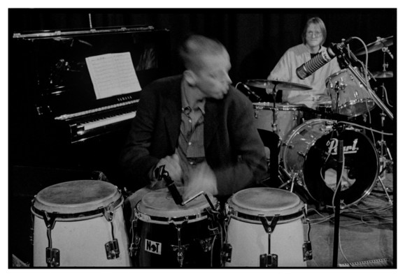 Black and white photograph of a percussionist with a drummer in the background. There is lots of motion blur on the percussionist's hands and face.