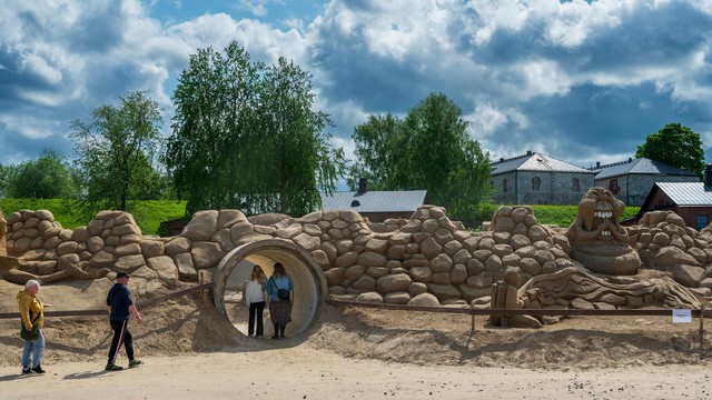 Hiekkalinna Lappeenranta. Valokuvaaja Kuvaajankulma Suomi.
Sandy terrain with a tunnel entrance surrounded by an artificial rocky structure. Two people standing inside the tunnel, while others walk nearby. On the right, a sand sculpture of a creature with a wide mouth and large eyes. In the background, green trees and stone buildings under a cloudy sky.