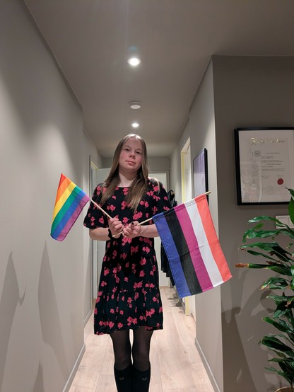 A person stands in a hallway holding a rainbow flag and a genderfluid pride flag, with a framed certificate and potted plant in the background.