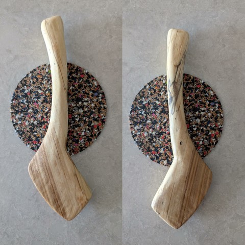 A collage of two pictures of a wooden butter spreader showing both sides. The piece is on a coaster for scale, on a grey stone countertop. Black lines of spalting have spread through the cream-coloured wood giving it character, the darker heartwood makes up part of the blade.