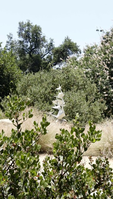 A white, modern sculpture tower of origami birds surrounded by green trees and bushes.