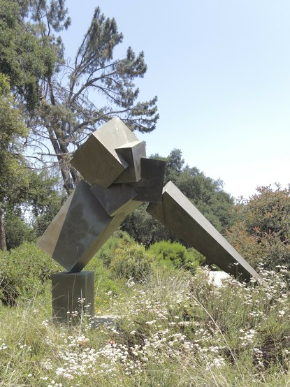 A geometric, metal modern sculpture surrounded by grasses, trees, and white flowers.