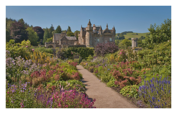 Large flower borders line a path leading to a large building in the distance. It's a sunny day with clear blue skies and the image is bright and colourful.