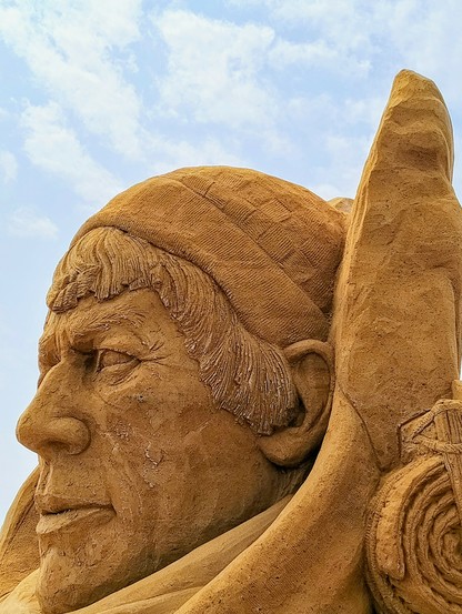A sand sculpture showing a head of an older man with weathered features. It's extremely realistic and detailed. The structure of the hat he's wearing looks like entrelac knitting.