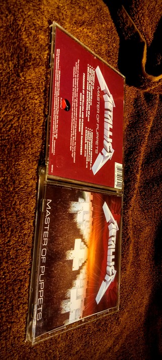 The photograph warmed with an 81A filter & bathed in 85F warm light, shows a CD case of the album "Master of Puppets" by Metallica. The case is open, revealing the back cover and the front cover. The back cover is red with white text, displaying the album title "Master of Puppets" in large white letters, along with the band name "Metallica" in a stylized font. The text includes production credits, track listing, and a copyright notice. The front cover features a dark background with a graphic of white crosses against a fiery sky, with the album title "Master of Puppets" in white text on the left side. The CD case is placed on a brown carpet, and a person's foot is visible in the top right corner of the image{?}

 Ovis2-8B

🌱 Energy used: 0.196 Wh