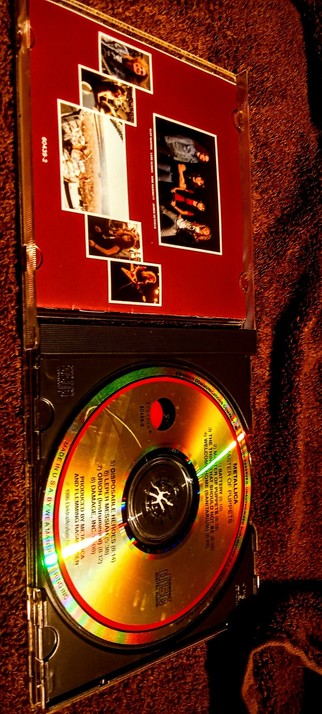 The photograph warmed with 85F gelled light and a 81A filter on the camera, shows a CD case with a red insert and a gold CD. The insert features a collage of five photographs, including images of people and a group, with a large central photo of a group of people. The CD has a reflective gold surface with a red ring around the center, displaying track titles and durations. The text on the CD includes "DISPOSABLE HEROES" and "METALLICA," along with other track names such as "WELCOME HOME (SANITARIUM)" and "LEPER MESSENGER." The case is open, revealing the CD and the insert. The background is a textured brown surface, possibly a carpet. The CD case is labeled "MADE IN U.S.A. BY WEAVER AUDIO PRICING" and "PRODUCED BY METALLICA AND FLEMMING RASMUSSEN."

 Ovis2-8B

🌱 Energy used: 0.214 Wh