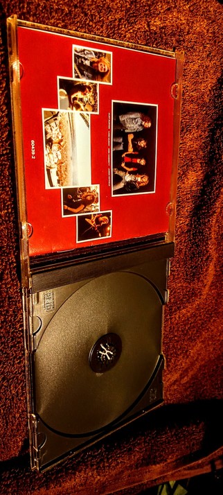 The photograph shows a CD case with a red background and several photographs. The case is open, revealing a black CD with a central logo. The top part of the case features a collage of six smaller images, including a person with long hair, a group of people, and a band performing. The bottom part of the case contains the CD, which is black with a central logo. The CD case is labeled with the number "60439-2" on the left side. The background is a textured, reddish-brown surface, possibly a carpet or fabric.

 Ovis2-8B

🌱 Energy used: 0.162 Wh