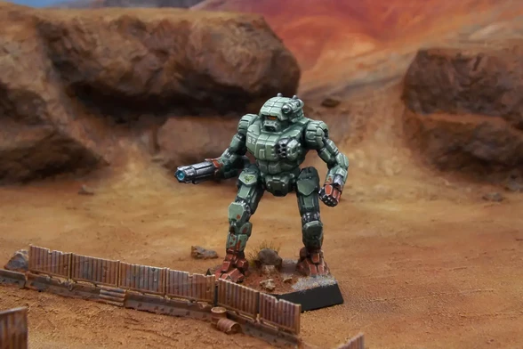 A painted miniature of a Vindicator 'Mech.  It is light green with a red blood splatter pattern on the forearms and feet.  The cockpit glass is orange.  The emblem of the Capellan Confederation is on the right leg, and the number "52" is on the right torso.  The miniature is photographed on desert terrain.