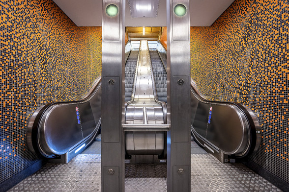 Welcoming escalator at Halemweg U-Bahn station in Berlin. The chosen angle and symmetry create a pareidolia effect showing a smiling robot with green eyes and open arms on orange and black mosaic background.