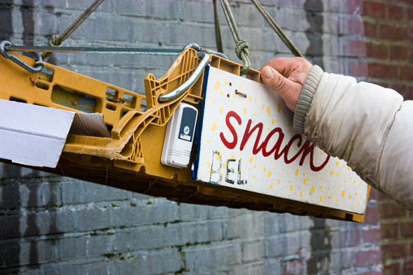 A yellow plastic crate hangs from a rope along the quay of a canal in The Hague. On it is a cardboard sign that says "Snack" and a sign that says "bell". A small walkie-talkie hangs next to it. A hand holds the crate, and a man (he is standing in a boat along the quay, but you can't see that) pulls it towards him. You only see his hand and arm.
