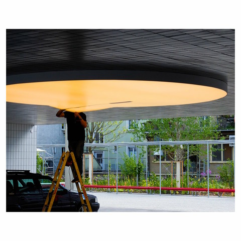 A man cleaning the inside of a huge yellowish ceiling lamp in a passageway. the man's head disappears in / behind the lamp.