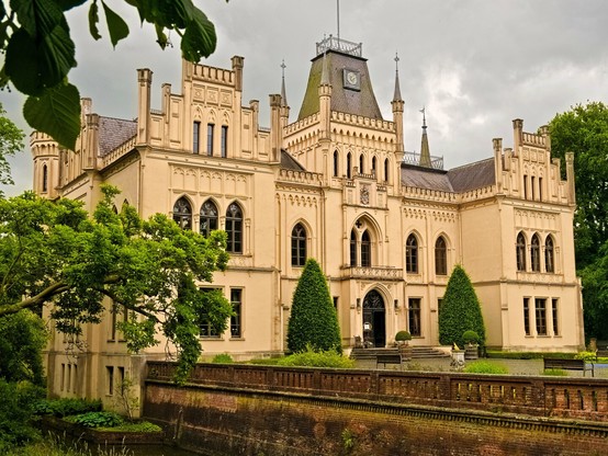 A yellowish moated castle in neo-Gothic style with battlements and arched windows against a very cloudy sky. In the foreground on the left, a tree juts into the picture. Even if it looks a bit like that, it's not a horror castle, I promise ;)