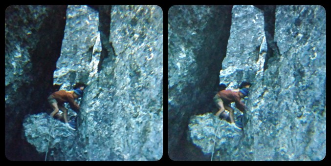We're looking at a rock climber in the middle distance. There are blue-gray rock walls on the right and left of us. The climber is on a small ledge, and is bent over, making adjustments to climbing gear, I assume. It's a 3D image, and there's kind of a keyhole effect behind the climber, with more rock surface behind them.