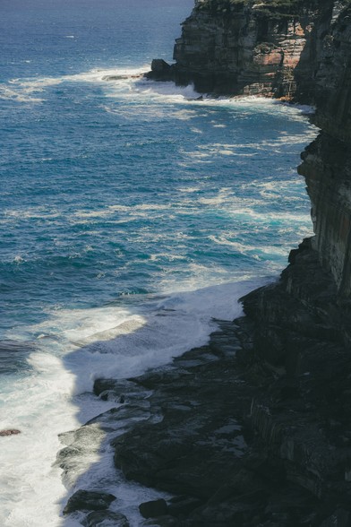 Rocky coastal cliff with waves crashing against the dark stone. The ocean displays varying shades of blue, illuminated by sunlight. A shadow is cast over part of the rocks, adding contrast to the scene.