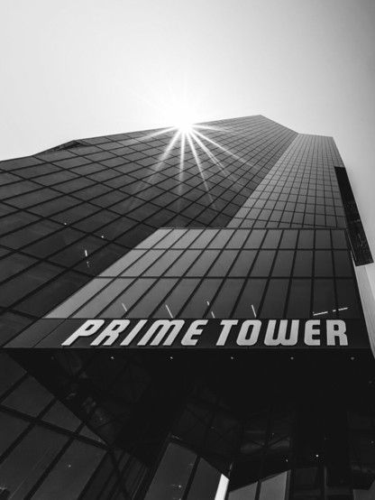 A dramatic upward view of the Prime Tower, a glass-clad skyscraper reflecting sunlight as it pierces through the top edge of the building. The bold white "PRIME TOWER" signage stands out against the dark façade in this high-contrast black-and-white image.