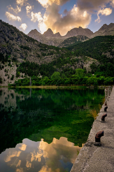 A vertical photograph showing a mountain landscape at sunset. In the foreground, a lake can be seen, and in the background, the Aragonese Pyrenees mountains, with the sun already behind them.