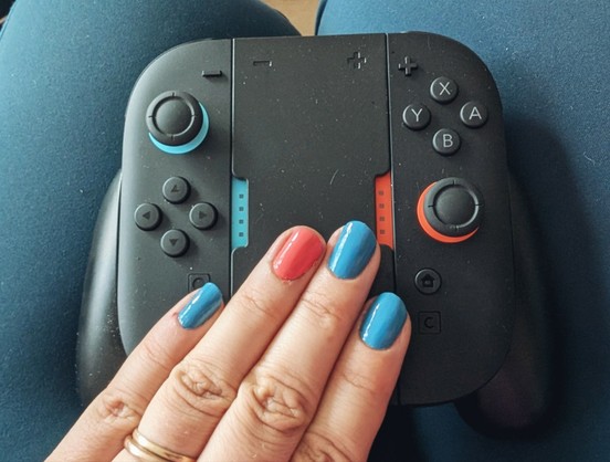 Ein Controller aus zwei Switch 2 Joycons und einem Verbindungsstück liegt auf einem Schoß, darauf eine Hand mit lackierten Nägeln und zwei Ringen.
Der Ringfinger ist in einem Korallenton lackiert, die anderen Finger hellblau, passend zu den Farbakzenten auf den ansonsten Schwarzen Controllern.