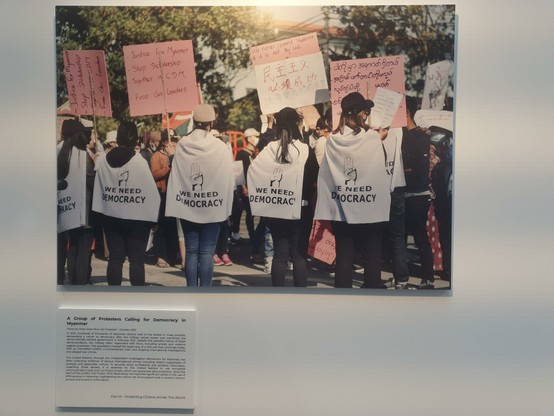 Photo of a group of protestors for democracy in Myanmar, showing people from the back wearing white capes that read "we need democracy".