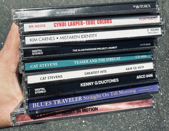 A hand holding a stack of CDs, including titles by Cyndi Lauper, Kim Carnes, Cat Stevens, Kenny G, Blues Traveler, and Amy Grant, on a pavement surface.