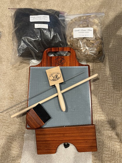 Top: two bags of good clean fiber, one merino and CVM and the other Corriedale. Middle, blending board made with paduak wood. On top of a board, a cleaning brush, wooden and metal dowels, and a blending brush. Bottom, keel for the blending board.