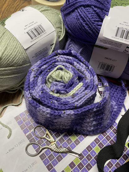 The central focus is a rolled strip of crochet in two shades of purple with a splash of a pale green. The larger skeins of yarn and the chart with numbers for the pattern are in the background.
