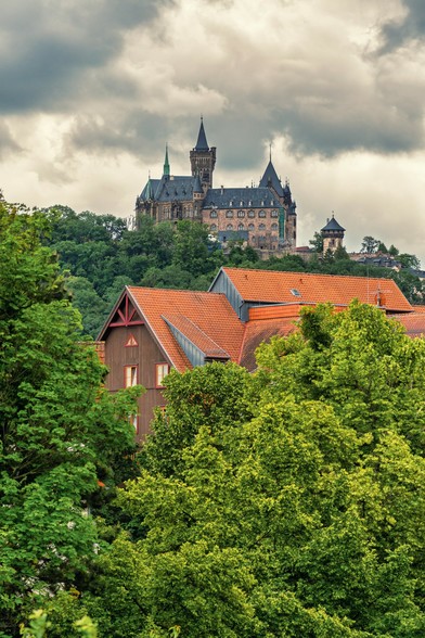 Im Vordergrund Dächer eines Gebäudes. Dahinter Wald. Im Hintergrund auf dem Berg das Wernigerode Schloß und darüber ein Wolkenhimmel kurz vor dem Regen