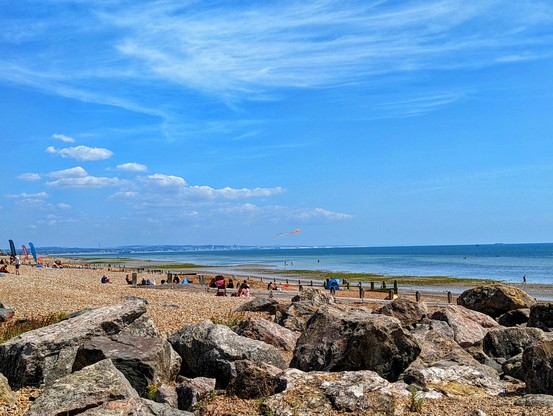 A panoramic view of Worthing beach on a sunny day. In the foreground, large, grey, weathered rocks are scattered across the shingle beach. Beyond the rocks, the beach transitions to smaller pebbles where several individuals are spread out, relaxing and enjoying the sunshine. Towards the horizon, the calm, blue sea stretches out, meeting the lighter blue sky with scattered white clouds. A long, faint landmass is visible in the distance across the water. A colourful kite with an orange tail is flying high in the sky. To the left, a few beach flags or banners are visible, adding splashes of colour to the scene. The overall impression is one of a peaceful, pleasant British seaside day.
