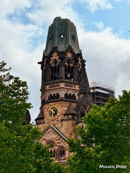 Blick auf den beschädigten Turm der Kaiser-Wilhelm-Gedächtniskirche in Berlin. Die Spitze ist durch den Zweiten Weltkrieg zerstört, das Mauerwerk wirkt gealtert. Zwischen den grünen Bäumen am unteren Bildrand hebt sich der rissige Turm mit seiner goldenen Uhr eindrucksvoll vom wolkigen Himmel ab.