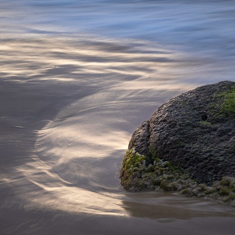 a rock looking like a stonefish at the beach with water flowing around it. The water is reflecting sunset light and making a curved shape on one side of the rock.