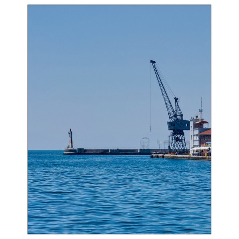 The image shows a serene coastal scene with a calm sea under a clear blue sky. On the left, a small lighthouse stands at the end of a pier. To the right, a large industrial crane is visible, likely part of a port facility. In the background, there is a spherical metal structure, adding to the industrial feel of the scene.