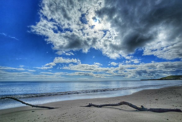 A wide shot of a serene beach scene with a bright blue sky partially obscured by dramatic, fluffy white and grey clouds. The sun is visible peeking through a break in the clouds. Gentle waves roll onto a sandy shore where two pieces of dark, gnarled driftwood lie in the foreground. The calm ocean stretches to the horizon, and in the distance to the right, a faint headland or pier is visible. The overall mood is peaceful and expansive.