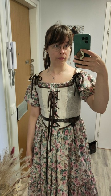 A mirror selfie of a white woman in an off-shoulder floral dress with an off-white and brown corset on top of the dress and a dark brown leather belt on top of the corset on the waist.