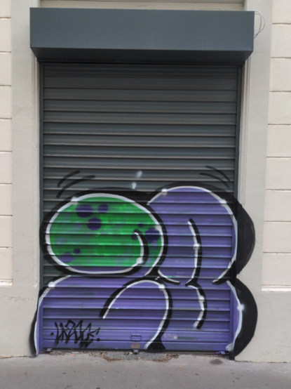Sur un rideau métallique vertical gris, un blaze en deux chiffres ronds violets "23" (?), le haut du "2" étant vert avec des points bleus, formant comme la tête d'un ver.