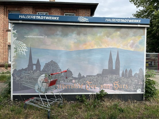 A shopping cart in front of a small transformer station (or something like that) featuring a slightly faded mural of a landscape with churches, greenery, and the text "Halberstadt: Tor zum Harz." (In Fractura). 

Photo: me