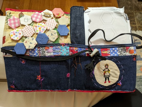 Fifteen hexagonal fabric patches with various printed patterns and motifs rest on a pieced fabric project folder which holds pattern paper and folded dark denim.