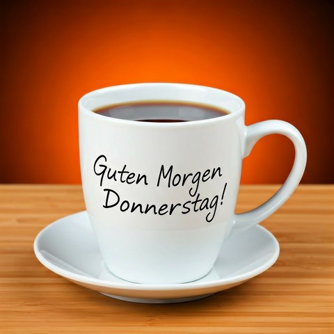 Auf dem Bild ist eine weiße Kaffeetasse mit Untertasse zu sehen, die auf einer Holzoberfläche steht. Die Tasse ist mit einem dunklen Getränk gefüllt, wahrscheinlich Kaffee. Auf der Tasse steht in schwarzer Schrift "Guten Morgen Donnerstag!". Der Hintergrund ist unscharf und orange-braun.