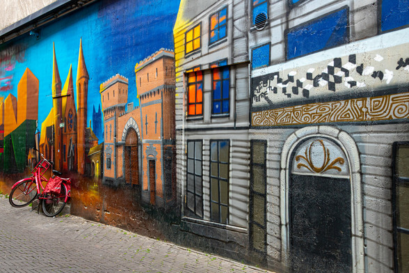 A colorful mural. The wall goes from right to left. At the beginning a gray house with doors and windows, then an orange castle and at the end a red fantasy castle with high red pointed towers. Above it is a blue sky. Against the red tower at the end stands a red painted bicycle, a real one. Bottom left in a slanted point of the street.