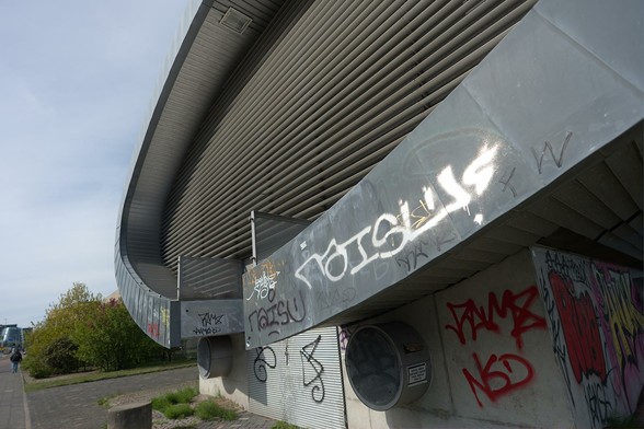 A large oval grey structure made of metal overhanging on a concrete foundation, with a facade resembling a huge grill. The structure is spray-painted with illegible street signatures. There's a cylinder sticking out at the base of the concrete part, with a sticker on it.
