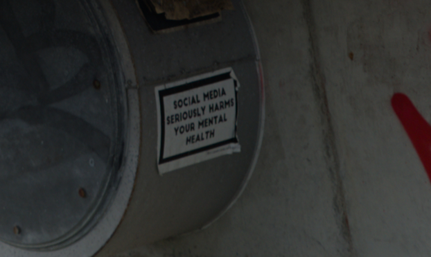 The sticker says:

"SOCIAL MEDIA SERIOUSLY HARMS YOUR MENTAL HEALTH"