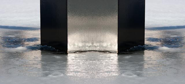 In a frozen pond, in the middle, stands a broad, black column, which reaches from the center of the photo to the top. The sun reflects in the shiny column. The ice is wet and the object reflects in it. About two-thirds of the way through the photo, the ice is dry and white snow-covered.
