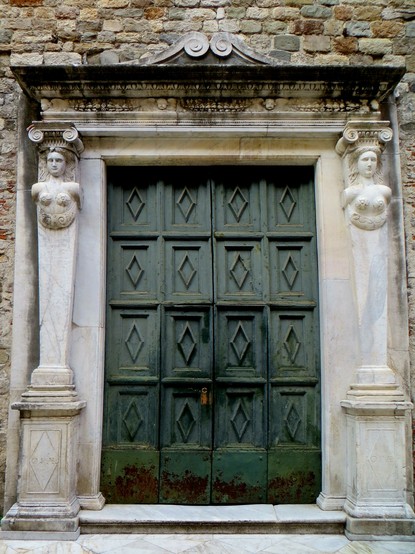 Picture of a church portal, with a green wooden door surrounded by a stone frame featuring two caryatids.