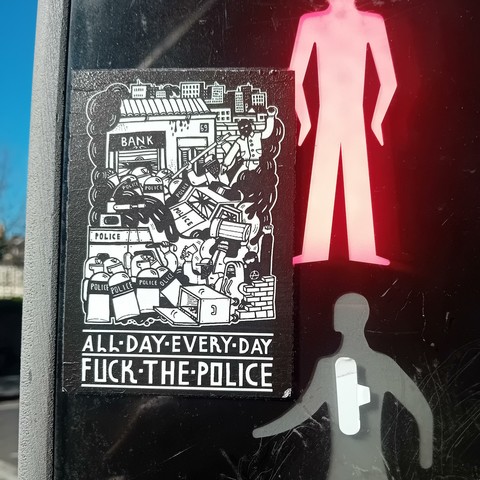 The image shows a sticker placed on a pedestrian crossing signal. The sticker is black and white and depicts a scene of unrest with police officers, riot shields, and a burning bank. Text at the bottom of the sticker reads:

ALL DAY · EVERY · DAY
FUCK THE POLICE