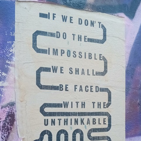 The image shows a poster that has been pasted on a street wall with a background of purple and blue graffiti. The poster features text are arranged in a staggered, zigzag pattern. The text reads: 

IF WE DON'T
DO THE IMPOSSIBLE
WE SHALL BE FACED
WITH THE UNTHINKABLE