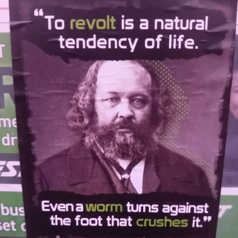 The image features a poster with a quote from and black-and-white portrait of Mikhail Bakunin, an anarchist philosopher and revolutionary. The quote reads:

"To revolt is a natural tendency of life.
Even a worm turns against the foot that crushes it."

Certain words ("revolt," "worm," and "crushes") are highlighted in green. The background is dark, and the overall design emphasizes the rebellious message.