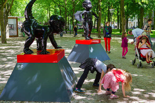 On the left of the photo is a statue of a black baby devil (a baby as big as a toddler) with horns on hands and feet and with tail and bottom up, on a gray pedestal with a red border. On the right of the photo. two little girls who, facing the statue, assume the same position, so on hands and feet with their bottoms up. One looks back to check if they are doing it right. All this on a path between trees, where some other visitors are also walking.